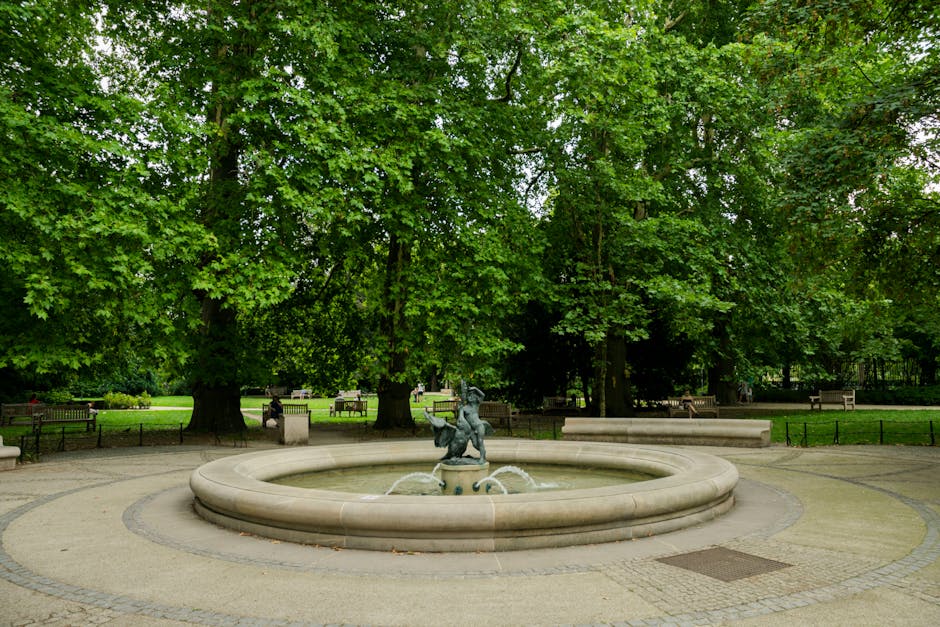 A serene view of a residential area overlooking a calm river with lush trees and well-maintained lawns. In the background, there are modern brick buildings with large windows, some with balconies. The