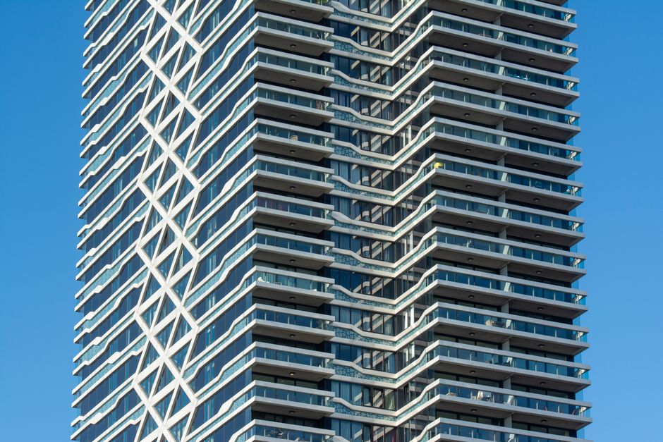 A low-angle view of a modern high-rise building with numerous glass windows and protruding balconies, emphasizing the clean and reflective surfaces of the exterior. The building's facade features a co