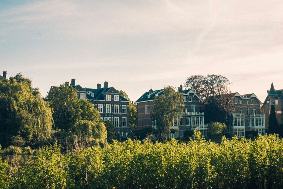 A residential area in Holland Park featuring a tranquil river reflecting the surrounding multi-story brick and stone apartment buildings with classical architectural details. The buildings have large