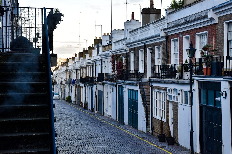 A row of residential houses on a sloped cobblestone street in Holland Park, London, with white and brick facades, black wrought iron railings, and potted plants. The image captures the exterior surfac
